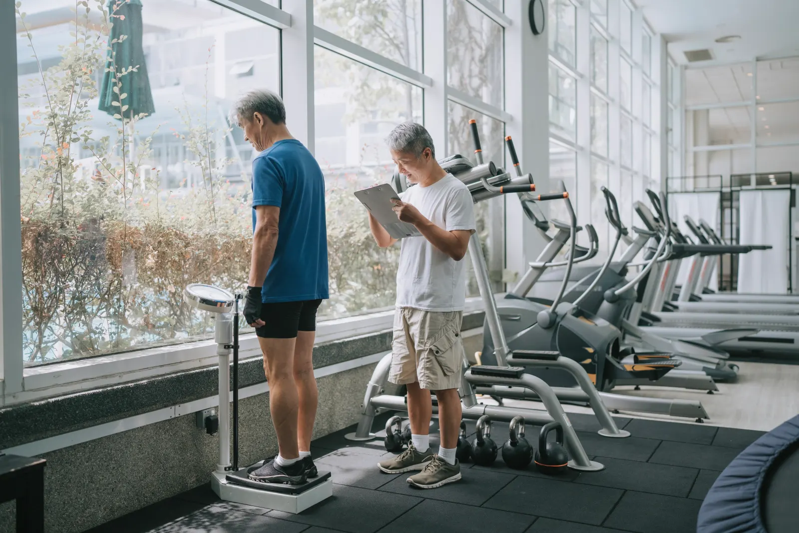 Trainer reviewing a chart as a man steps on a gym scale during a weight loss progress assessment