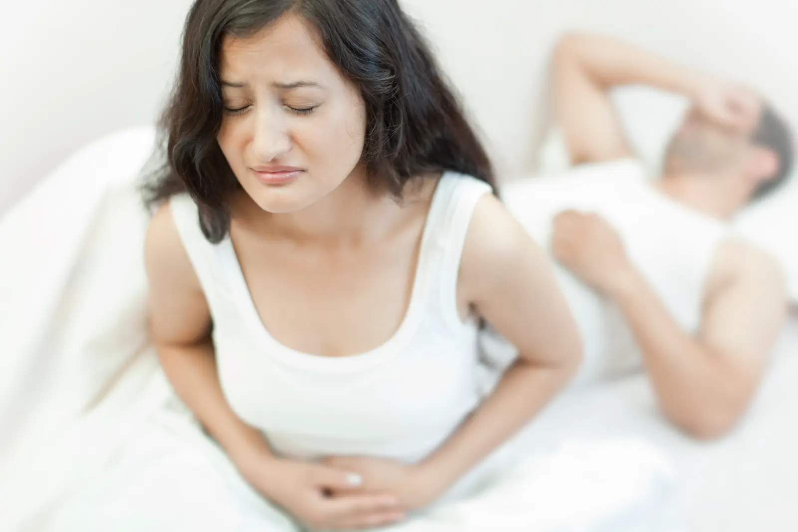 Woman clutching her stomach in bed beside a sleeping man, showing hangover nausea and discomfort