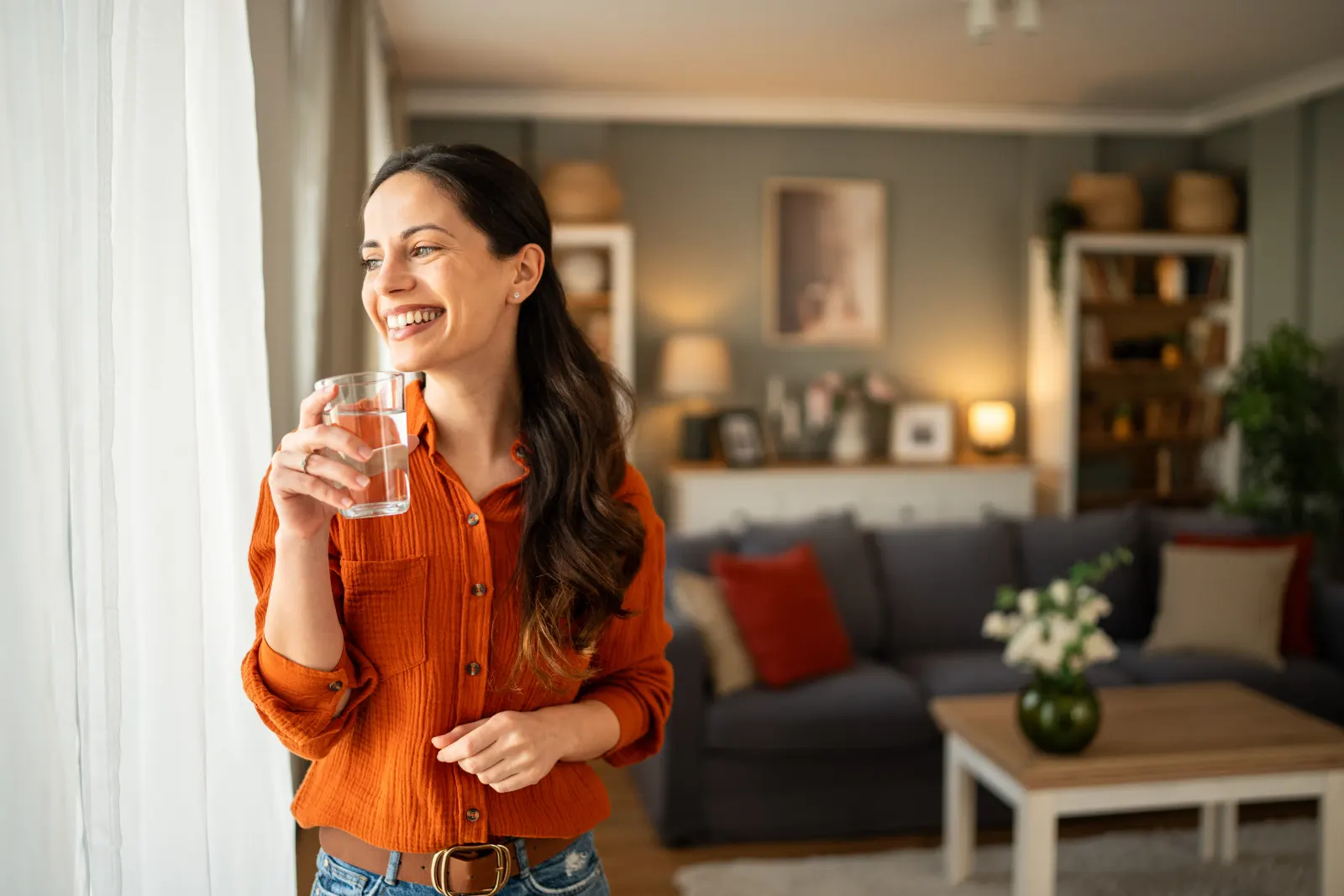 Woman in an orange shirt smiling by a window holding a glass of water at home in Westchester County